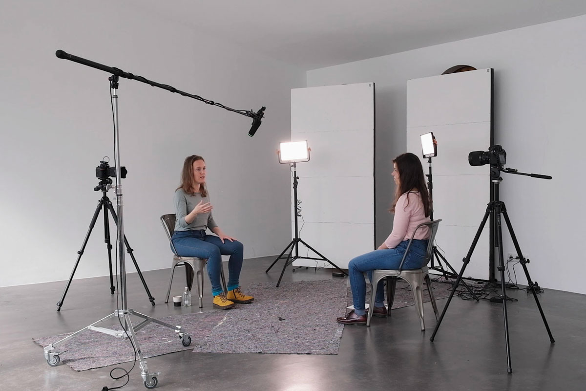 Auf dem Foto sind zwei junge Frauen zu sehen, die sich in einer Aufnahmesituation mit Kamera, Mikrofon und Scheinwerfern auf Stühlen gegenüber sitzen