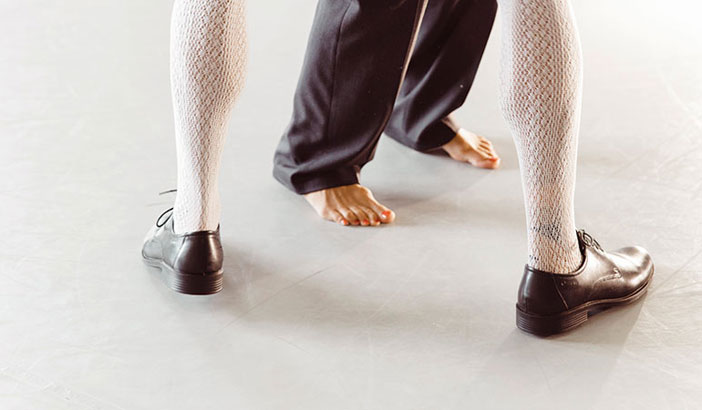 Zu sehen sind die Beine und Füße von zwei Personen. Eine ist mit weißen Spitzenstrümpfen und Herrenschuhen gekleidet, die andere trägt eine schwarze Hose keine Schuhe.