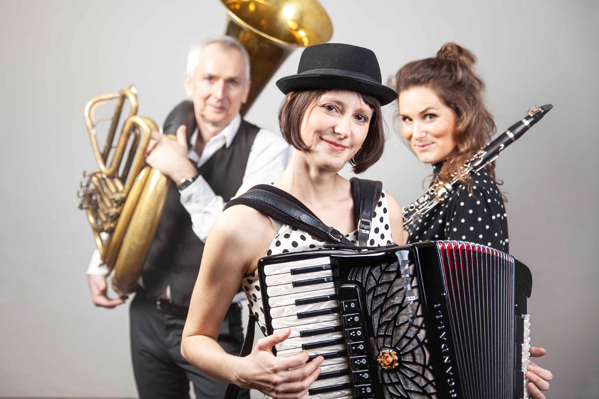 Eine Akkordeonspielerin steht im Vordergrund, im Hintergrund stehen eine Klarinettistin und ein Tubaspieler.